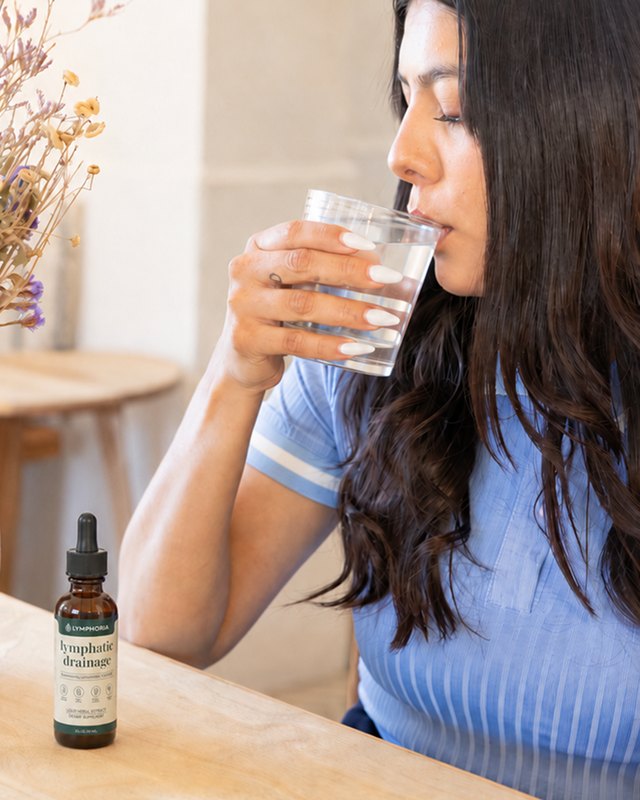 Una mujer bebiendo su vaso de agua, con el frasco de Lymphoria sobre la mesa